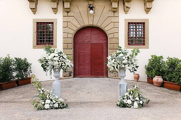 Ceremony entrance decor with wedding floral urns of white blooms and greenery flanking an arched doorway with red double doors in a courtyard