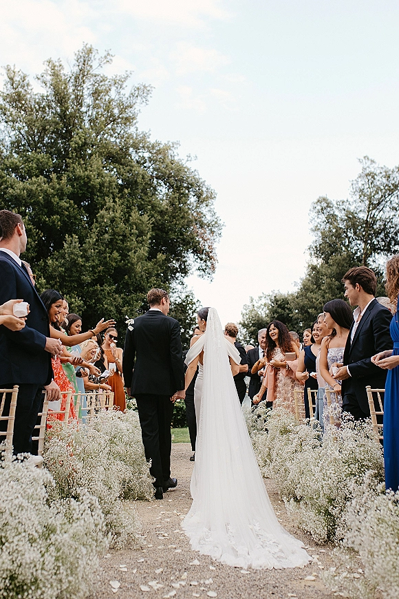 Wedding recessional as bride and groom walking away down an outdoor wedding aisle, guests cheering beside chiavari chairs in a garden