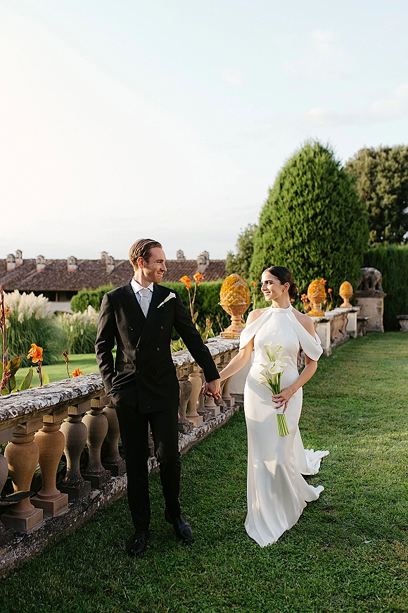 Couple portrait of bride and groom holding hands on a garden lawn, her calla lily bouquet and sleek dress beside his tuxedo by a stone balustrade