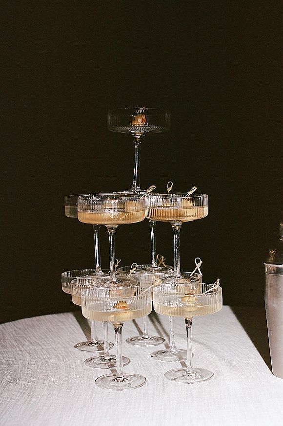 Champagne tower of stacked coupe glasses filled with bubbly, cocktail picks and warm candlelight on a white linen table against a dark wall