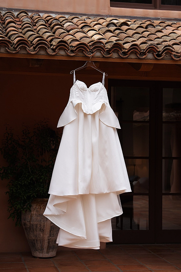 Wedding dress hanging on a hanger, showcasing spaghetti straps and corset bodice, with layered skirt against a stucco patio backdrop