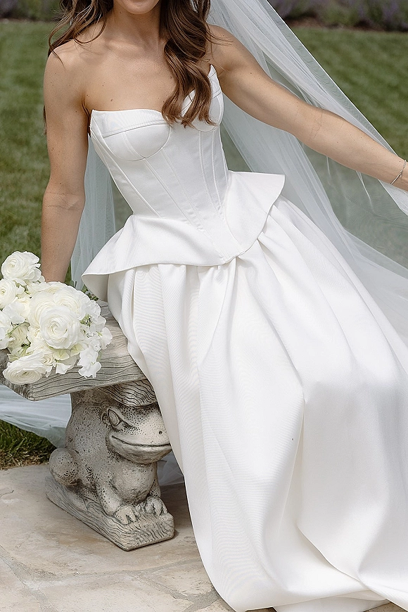 Wedding dress with a strapless wedding dress corset bodice, peplum waist, cathedral veil, and white rose bouquet on a stone patio lawn