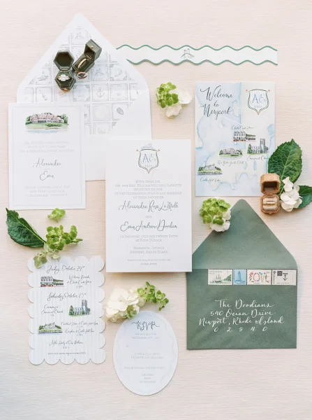 Invitation suite wedding invitations flat lay with green envelopes, wax seal, stamps, rings and map card on neutral fabric with greenery and white flowers