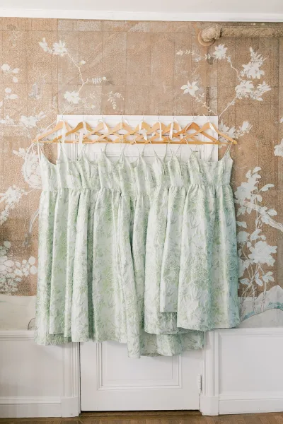 Bridesmaid dresses in sage green and floral print hang on a rack with wooden hangers against vintage floral wallpaper and a white door