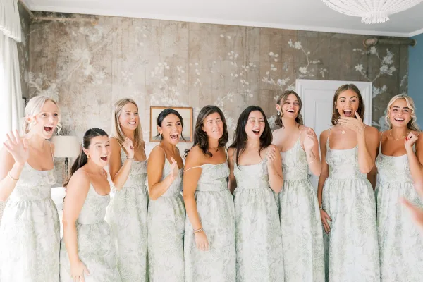 Bridesmaid group photo of bridesmaids getting ready, laughing in matching floral spaghetti-strap dresses by window light in a mural room