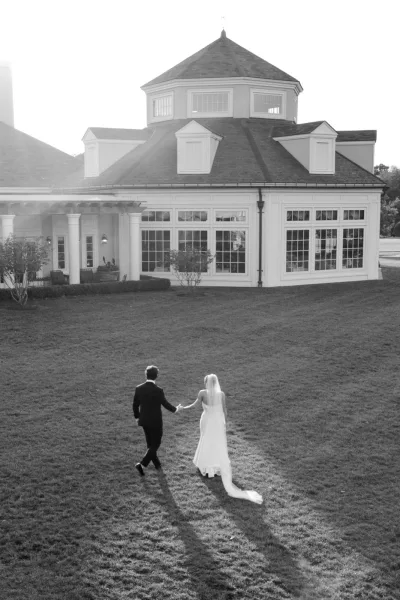 Bride and groom walking, holding hands across a grass lawn, her long veil trailing, toward a building facade with large windows and a columned porch.