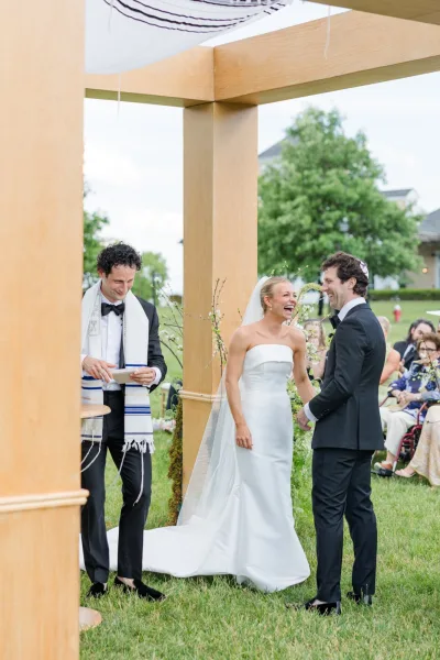 Ceremony moment at a chuppah ceremony as the bride laughs with the groom, hands clasped beneath the chuppah in a candid, joyful pause.