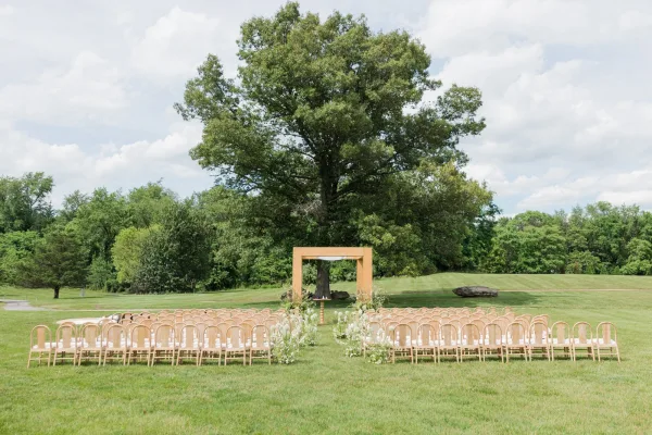 Outdoor ceremony setup with a wooden wedding arch draped in soft fabric on a grass lawn beneath a large tree and open sky.