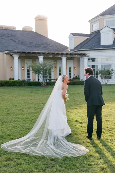 Couple portrait of a bride and groom laughing on a lawn, her veil visible, backed by a building with white columns and shrubs.