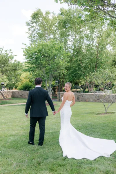 Couple portrait of bride and groom walking hand in hand along a garden path, her dress train trailing as they pass a low stone wall.