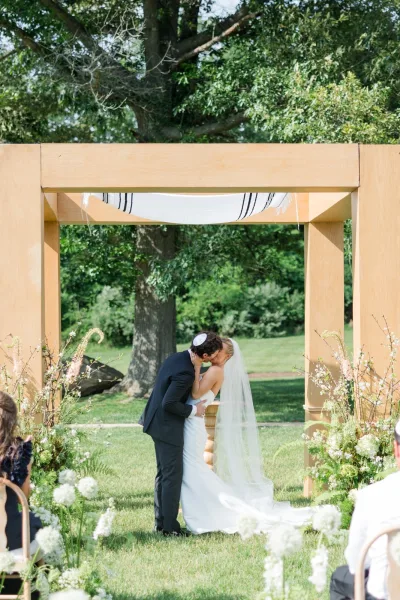 Ceremony kiss moment as the couple shares their “you may kiss the bride” under a wooden chuppah, framed by lush garden greenery.