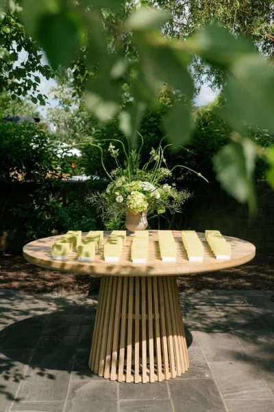 Escort card display serves as a place card display on a round wood table, framed by garden greenery and leafy branches on a stone patio.