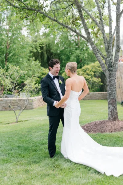 Wedding first look shows the groom seeing bride, his black bow tie crisp, by garden trees and a stone wall.