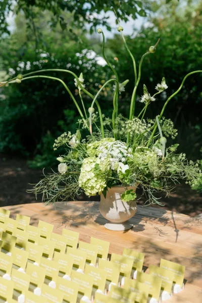 Escort card display with wedding place cards arranged neatly, anchored by hydrangea, against garden trees and a clear blue sky.