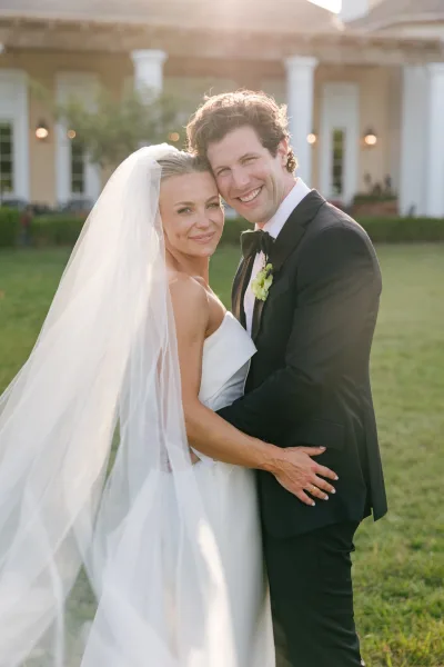Couple portrait of a bride and groom hugging, her long veil trailing as they embrace on a grass lawn before a building with white columns.