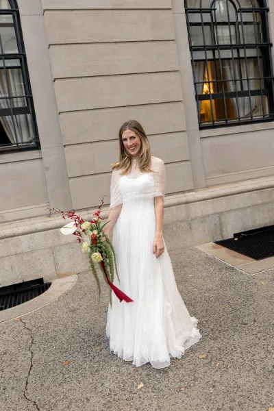 Bridal portrait of a bride holding a bouquet with a red ribbon, posed by a stone building facade with arched windows along a sidewalk.