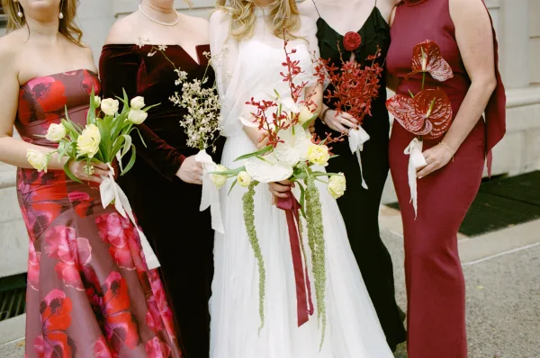 Bride and bridesmaids in mismatched bridesmaid dresses stand holding mixed bouquets with silk ribbons softly streaming.