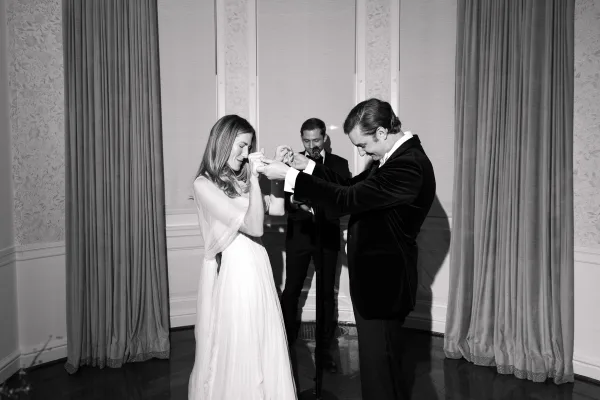 Ceremony moment exchanging vows as the bride and groom hold hands, with the officiant speaking into a microphone before indoor drapery.