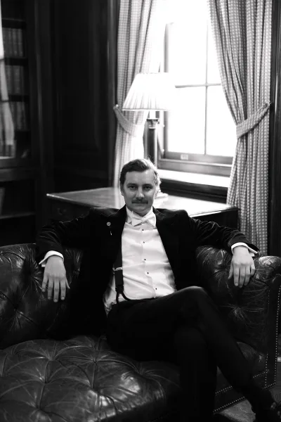 Groom portrait of a black tie groom seated on a leather sofa, framed by wood paneling and a bookshelf softly lit by a table lamp.