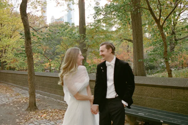 Couple portrait, an outdoor wedding portrait by a stone wall and autumn trees, the bride in a tulle wrap beside the groom.