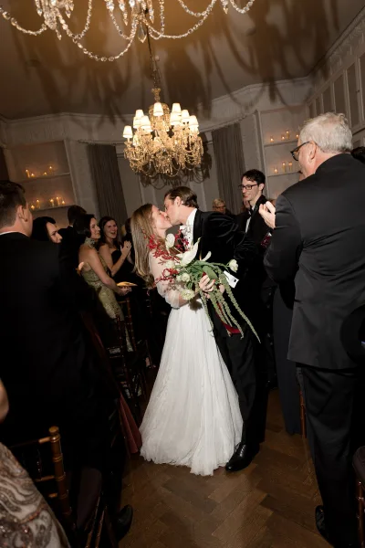 Wedding kiss moment during the ceremony recessional as the couple embrace on a candlelit aisle, guests applauding by drapery and paneled walls.