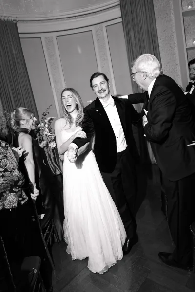 Ceremony recessional with bride and groom walking hand in hand, her bridal gown flowing, guests cheering in a draped ballroom with paneled walls.