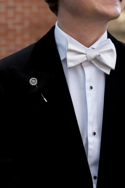 Groom bow tie showcased—the white bow tie rests neatly above a tuxedo’s velvet lapel, set against a textured brick wall.