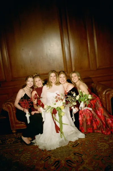 Bridal party portrait of a bride with bridesmaids in mismatched dresses, holding bouquets on a leather sofa by wood paneling.