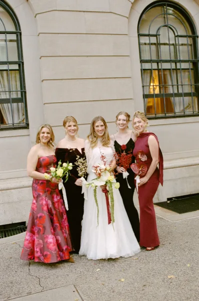 Bride and bridesmaids in mismatched bridesmaid dresses stand with bouquets by a stone wall and arched windows.