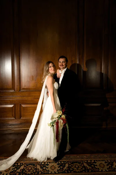 Couple portrait, an indoor wedding portrait with the bride holding a red and white bouquet beside the groom before a wood paneled wall.