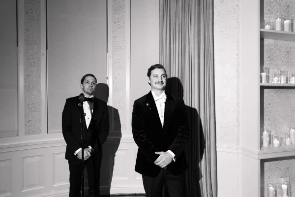 Groom portrait in a classic tuxedo with a white bow tie, smiling with hands clasped, set against indoor curtains and a paneled wall.