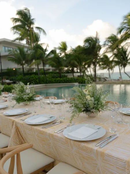 Reception tablescape styled for an outdoor reception table beside a pool with palm trees, featuring greenery centerpieces and ocean views.