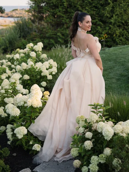 Bridal portrait of a bride in a strapless gown with tulle overskirt and ribbon choker, looking back on a stone garden path by hydrangeas