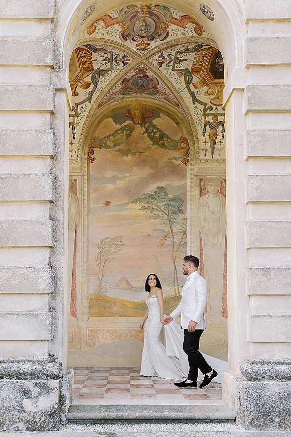 Couple portrait of bride and groom holding hands, her strapless gown with long train trailing on a checkered floor beneath a fresco archway