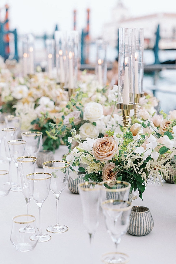 Reception tablescape with wedding table centerpiece of blush roses and greenery, taper candles in hurricanes, gold candlesticks, and waterfront pier view