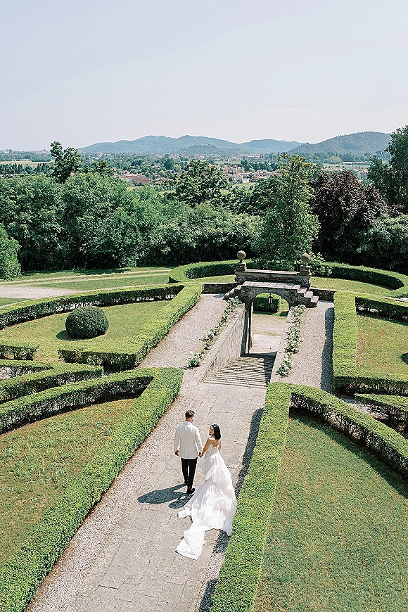 Couple portrait of bride and groom walking away hand in hand, her long train flowing, beneath a stone archway in a formal garden