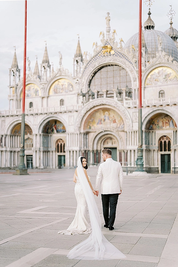 Couple portrait of bride and groom holding hands, walking away as the bride looks back, veil and train flowing before a domed cathedral facade