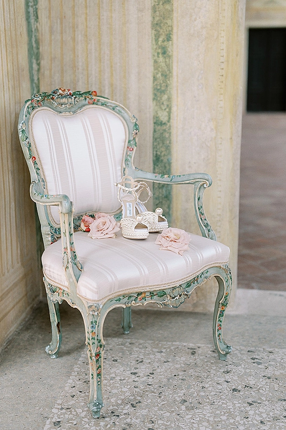 Bridal shoes with pearl wedding heels set beside pink roses on an upholstered vintage chair in a stone-column hallway with terrazzo floor
