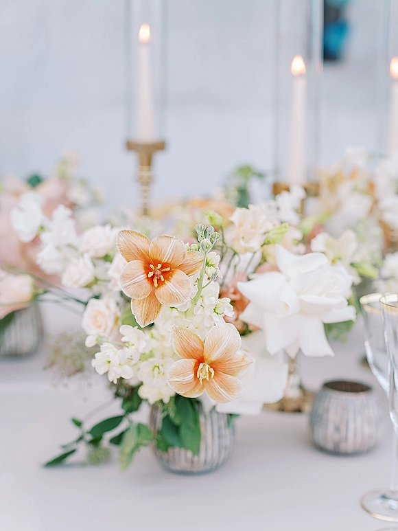 Reception centerpiece with wedding floral centerpiece of peach and white blooms in a glass vase, brass taper candles and votives on linens before a white paneled wall