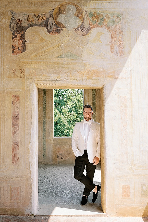 Groom portrait in a white tux jacket leaning in a sunlit arched doorway, stone walls and fresco mural behind, wristwatch visible