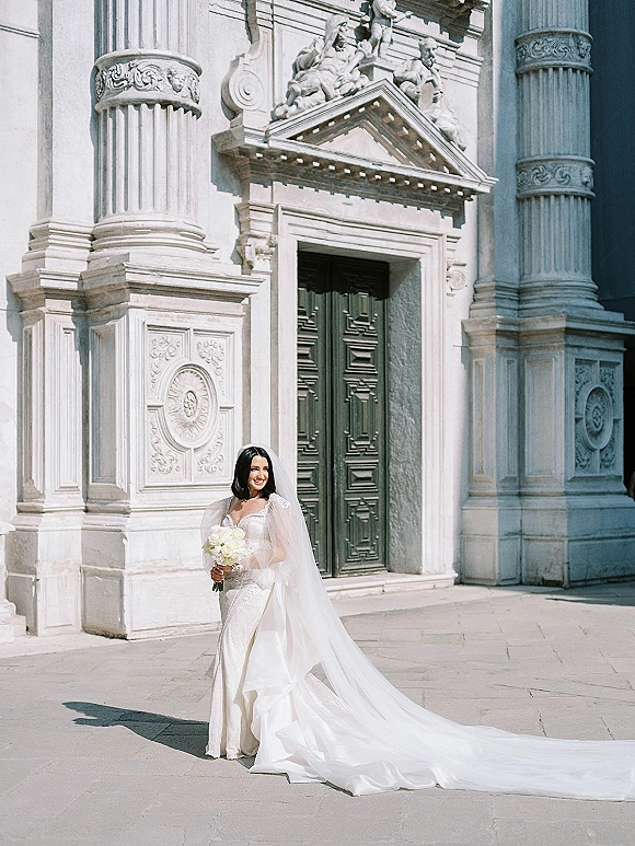 Bridal portrait of a bride in a wedding dress with long train and veil, holding a white bouquet before an ornate stone doorway in sunlight