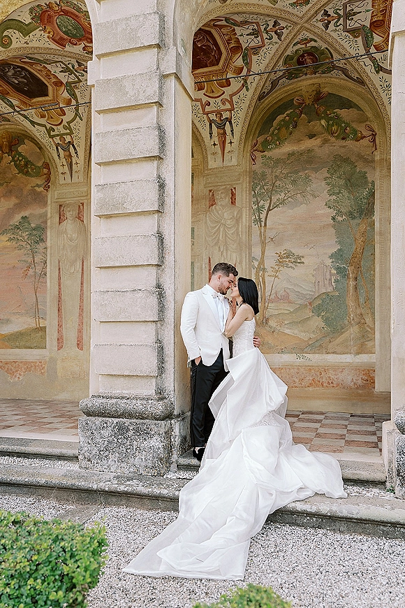 Wedding couple portrait of bride and groom kiss, her strapless gown’s long train flowing beside stone columns under a frescoed portico