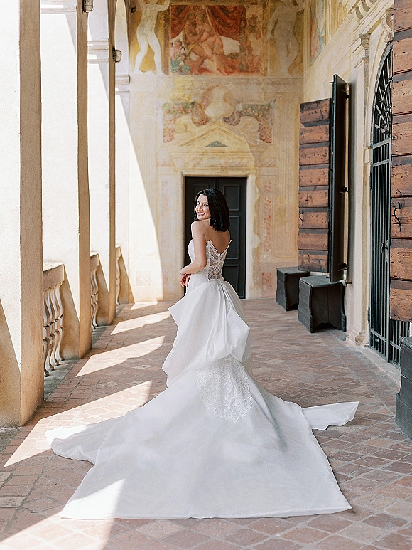 Bridal portrait of a bride looking back in a strapless gown, holding her wedding dress train in a sunlit arched corridor with fresco walls