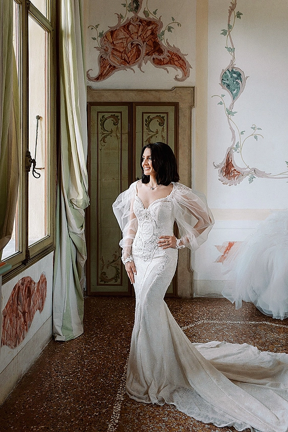 Bridal portrait of a bride by window in a lace wedding dress with puff sleeves and long train, softly lit in an ornate room with murals