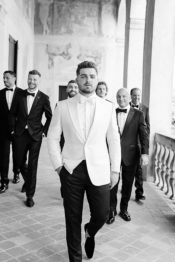 Groom portrait in a white dinner jacket and bow tie with boutonniere, standing in an arched corridor with columns and stone floor