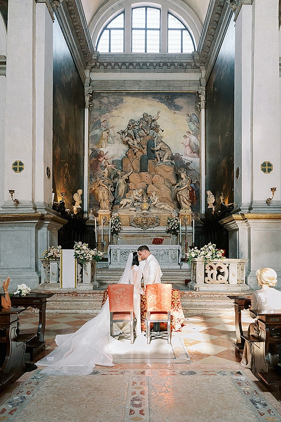 Ceremony kiss at a church wedding ceremony, bride in long train dress and veil with groom at floral altar and tall candles, arched window backdrop