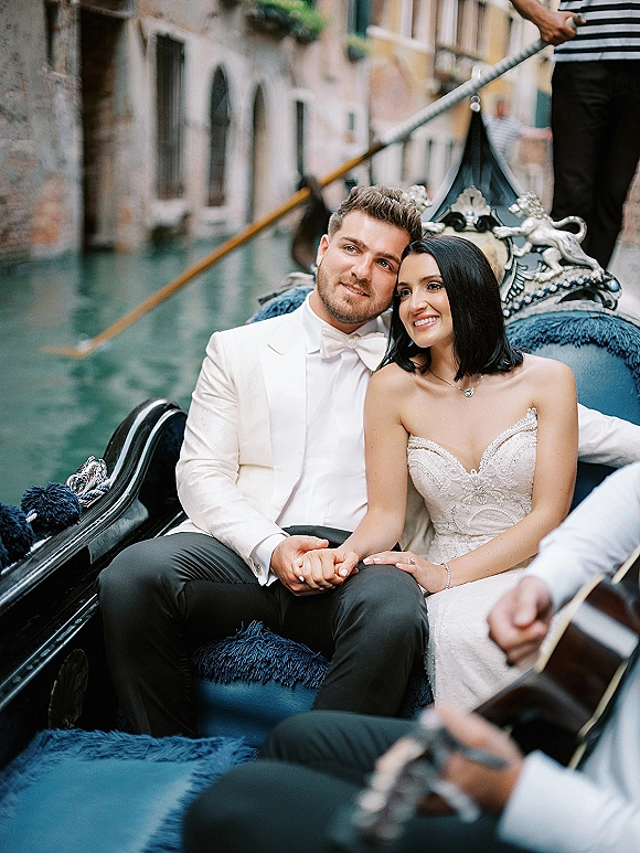 Couple portrait of bride in strapless lace gown and groom in white tuxedo holding hands on a gondola, canal buildings behind them