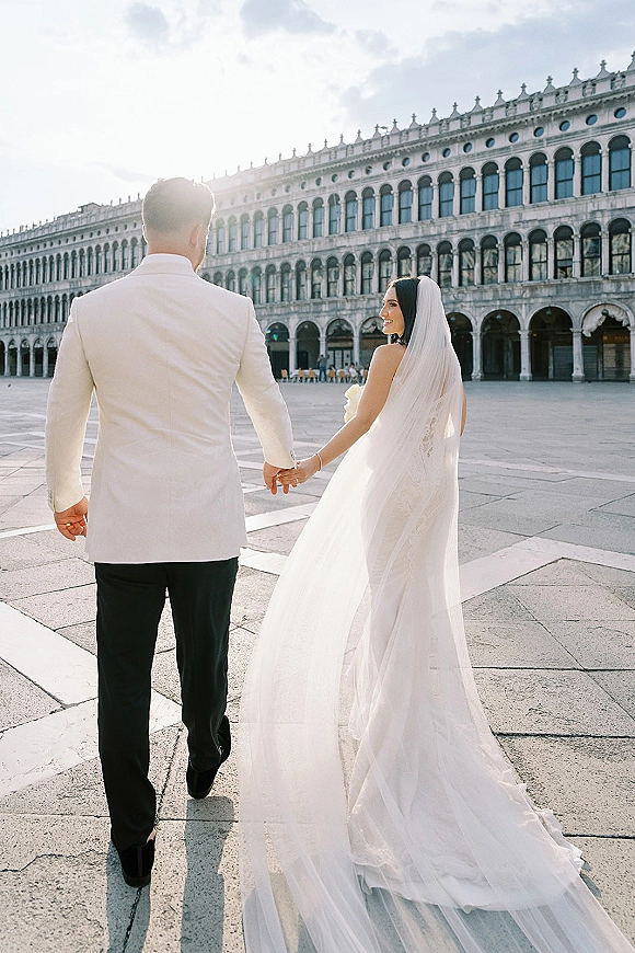 Couple portrait of bride and groom holding hands walking away, bride looking back as veil and train flow by a historic facade with sun flare