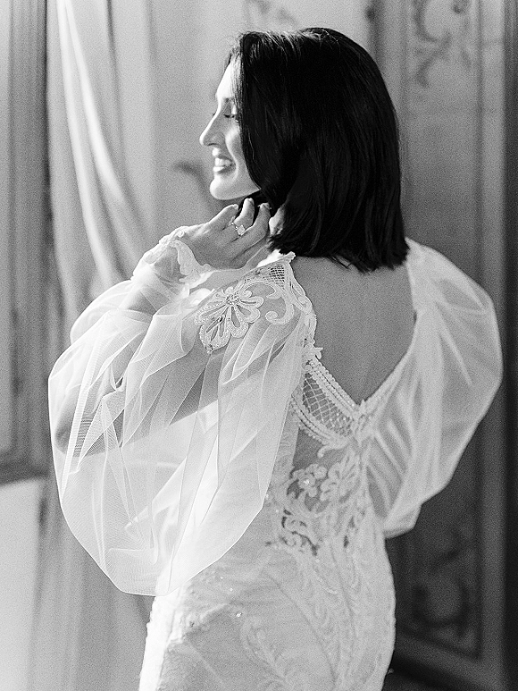 Bridal portrait in black and white of a smiling bride in profile adjusting her necklace, showing lace bodice and sheer puff sleeves by curtains