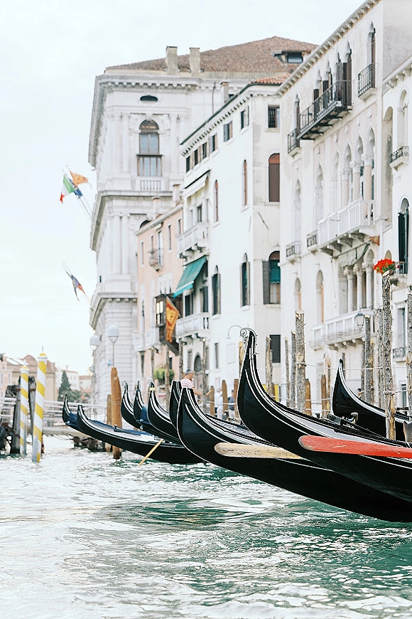 Gondolas on canal moored at a gondola dock scene, lined beside wooden poles with flags against historic waterfront buildings and balconies